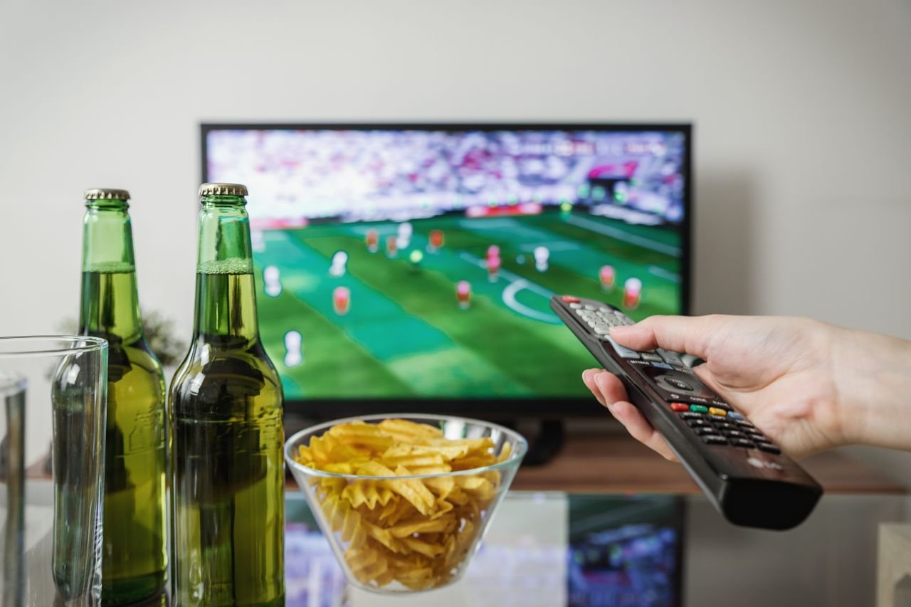 Home A person holding a remote while watching soccer on TV with beer and snacks on the table.
