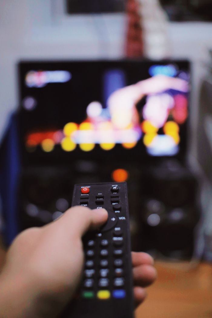 Crop unrecognizable person switching channels on remote control while watching TV on blurred background in light room during free time