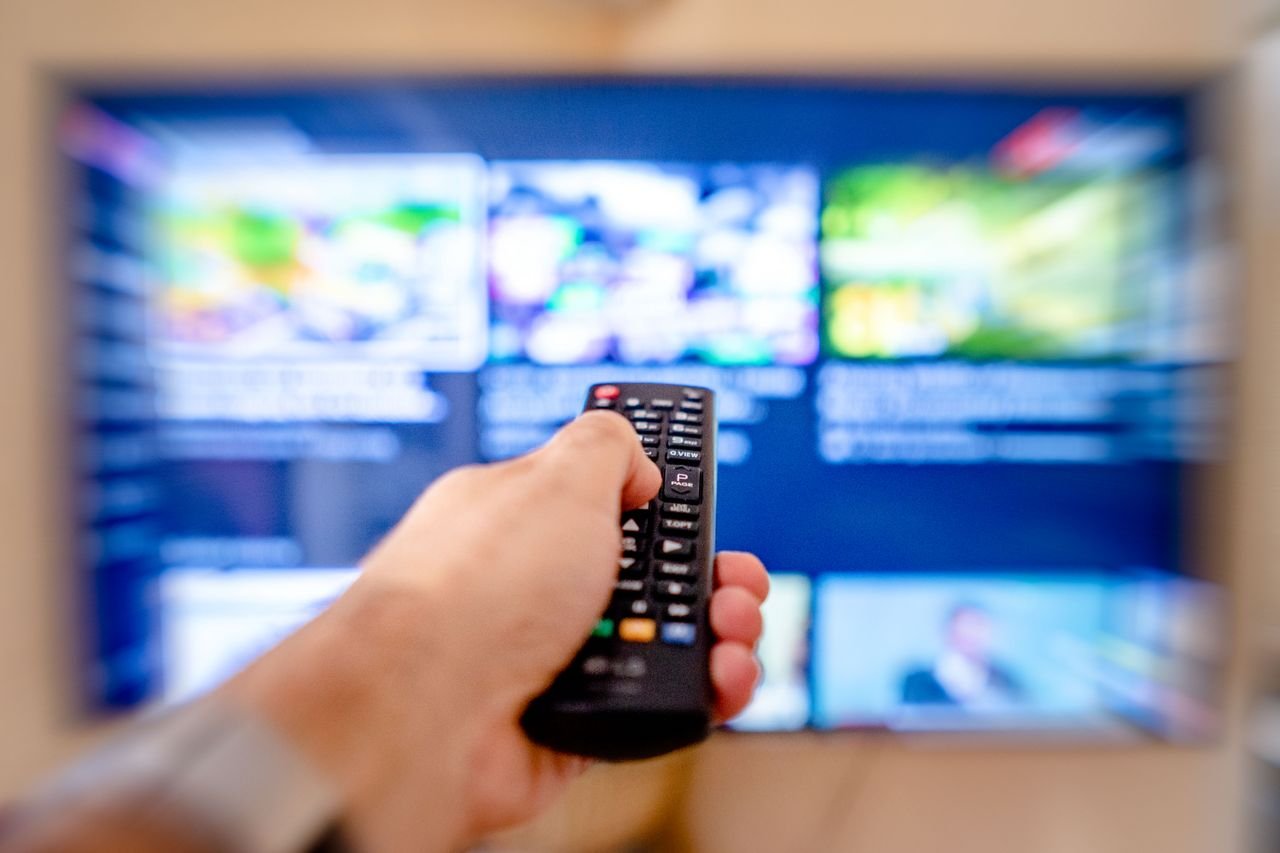 Hello world! Hand holding remote control pointing at a blurred TV screen in a cozy indoor setting.